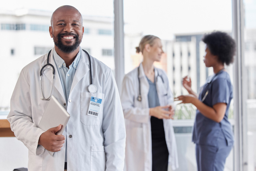 Financial planning for healthcare providers concept image with smiling doctor holding digital tablet and nurse and technician in discussion behind him.