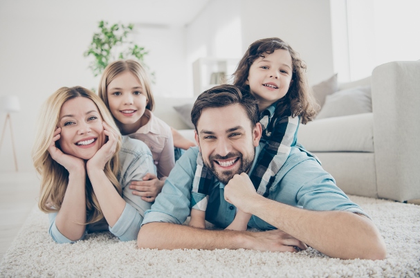 Kiddie tax concept depicted by photo of four members of family, mom, dad, and two young daughters on their backs, lying face down on floor and smiling at the camera