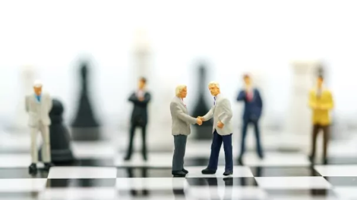 Business transfers is depicted through small figures of businessmen shaking hands while standing on a chess board.