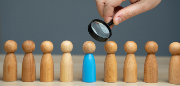 3P Planning Approach development step image concept has a man's hand holding a spyglass over a row of wooden pegs made to resemble people, and focused over one wooden peg that is painted blue.